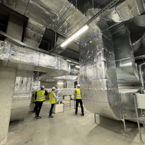 Visite d’un espace technique dans un bâtiment, où plusieurs personnes en gilets haute visibilité et casques de sécurité observent un réseau complexe de gaines de ventilation recouvertes d’isolant réfléchissant. L’ambiance industrielle est soulignée par les structures métalliques et les éclairages suspendus